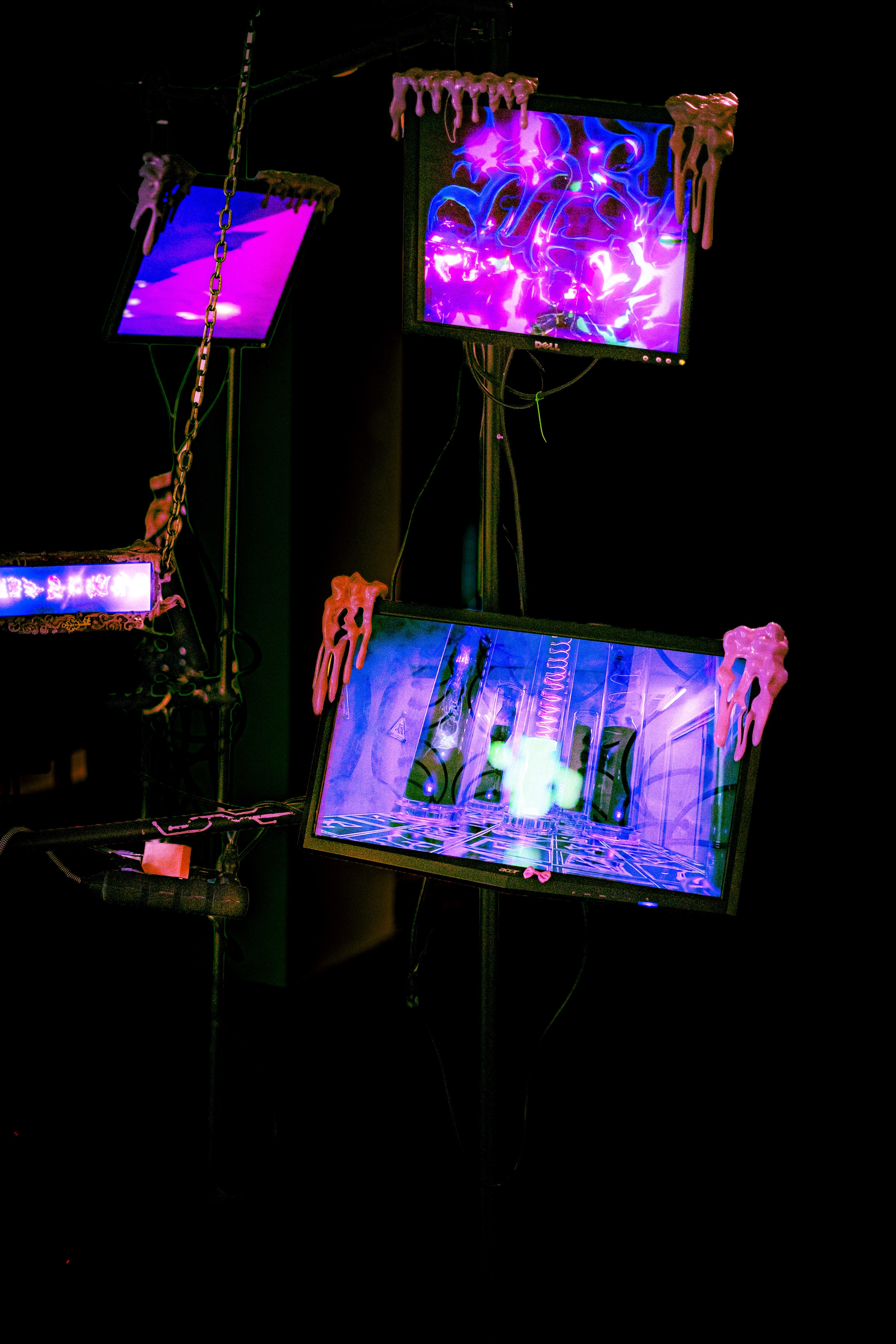 three stacked monitors on scaffold, pink slime clawing over their frames, the screens showing a chrome knot of violet light, a flat magenta field, and a long purple corridor with a glowing green cross down its centre, a slime-framed letterbox panel at far left