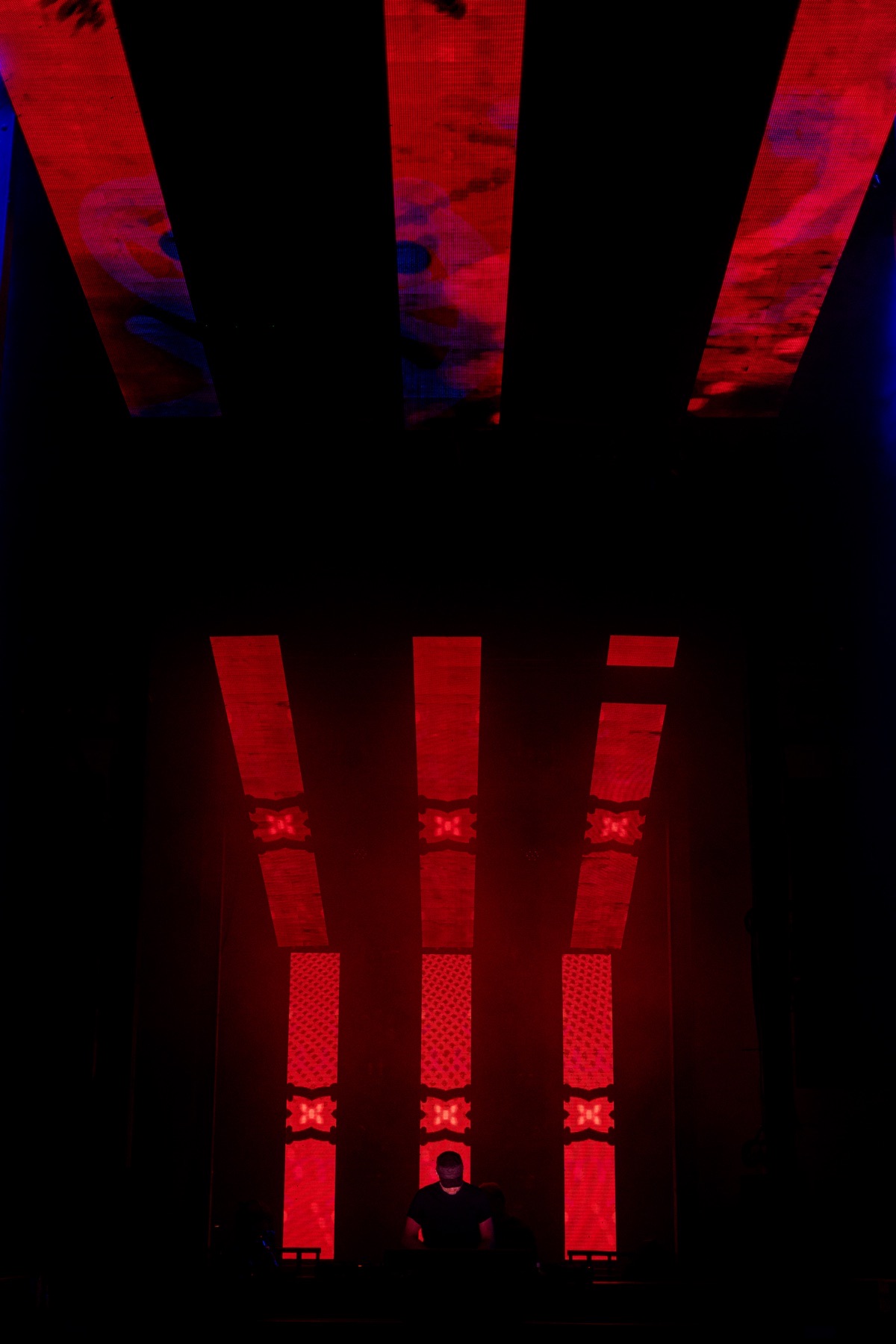 three panels glow red with mirrored x patterns, three more above them wash the ceiling in red and blue, a dj lit small at the very bottom between them.